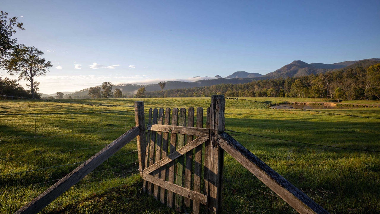 An image of a wire fence with a wooden gate. It is surrounded by a green landscape with distant hills.